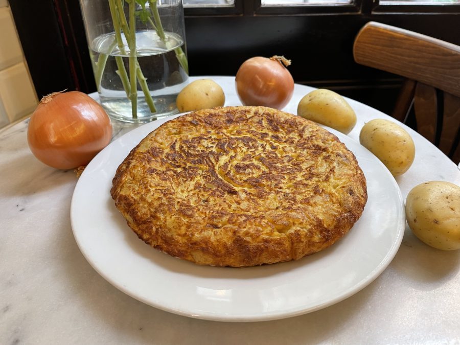 Tortilla de Patata y cebolla en un plato con ingredientes base a su alrededor, a la venta en el Obrador de la nena, Barcelona