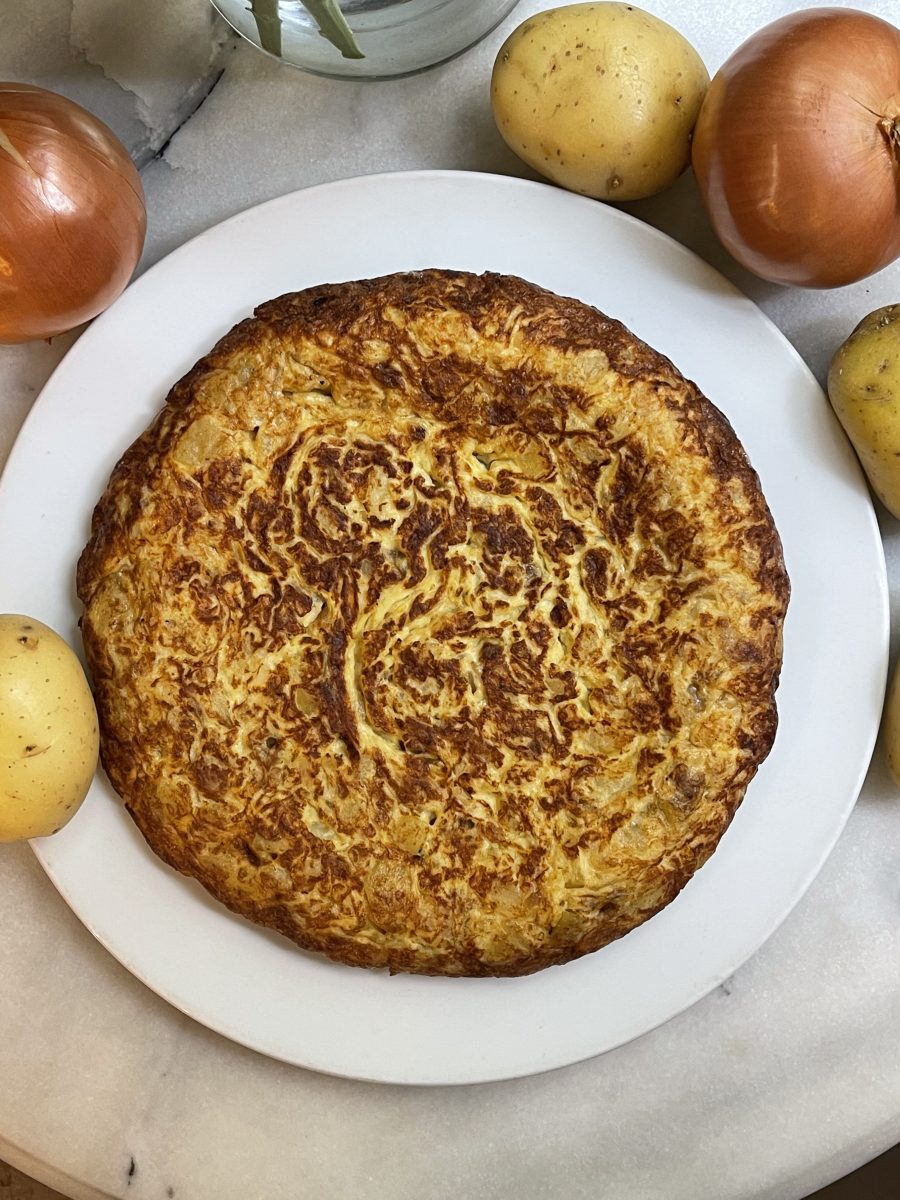 Tortilla de Patata y cebolla en un plato con ingredientes base a su alrededor, a la venta en el Obrador de la nena, Barcelona